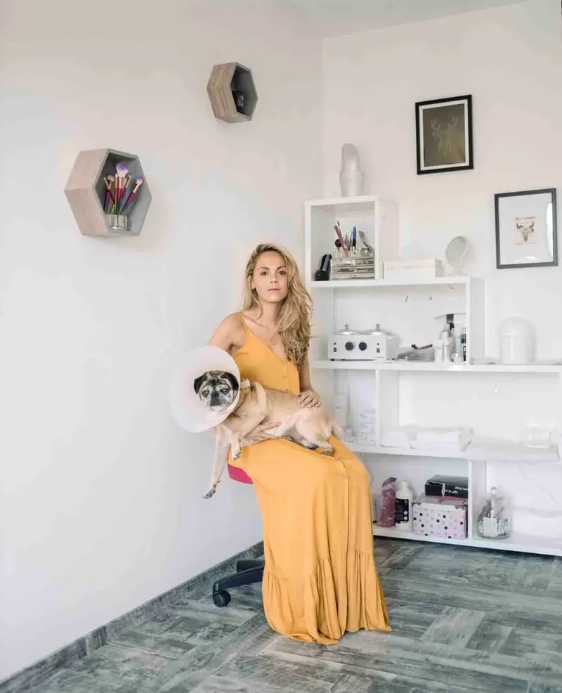 Portrait d'une femme blonde en robe jaune tenant un carlin à collerette, dans un intérieur blanc minimaliste aux accents esthétiques, éclairé de lumière diffuse naturelle