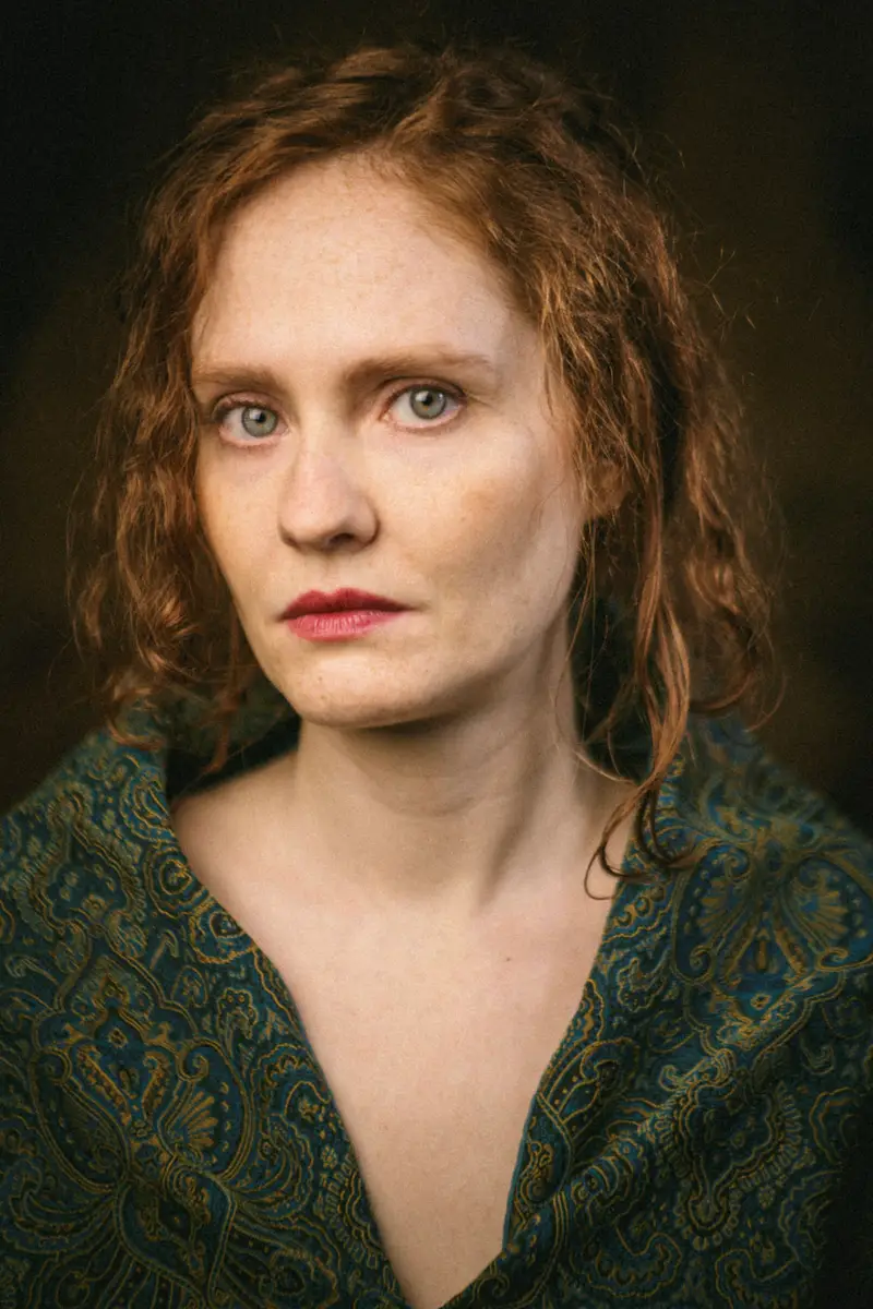 Portrait d'une femme rousse aux yeux clairs, vêtue d'un habit bleu-vert brodé d'or, éclairée par une lumière douce et dramatique dans un style pictural rappelant la Renaissance