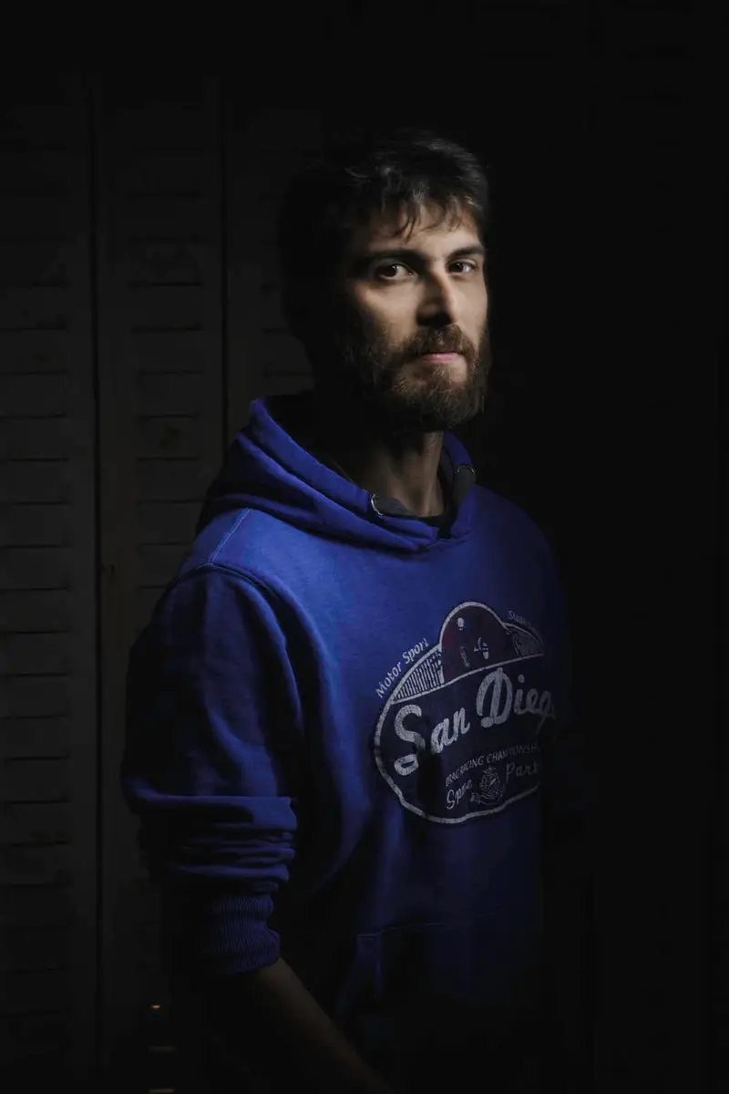 Portrait d'un homme barbu en sweat violet, éclairé latéralement dans un style chiaroscuro dramatique sur fond noir