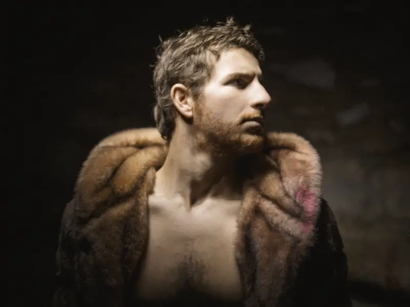 Portrait d'un jeune homme barbu au torse nu enveloppé d'un manteau de fourrure, baigné d'un clair-obscur dramatique dans un style pictural baroque