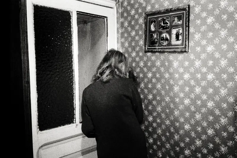 Femme de dos face à une porte vitrée, intérieur vintage au papier peint fleuri, cadre de portraits familiaux