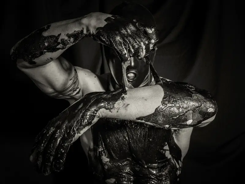 Portrait en noir et blanc d'une personne au visage masqué et au corps couvert de peinture noire visqueuse, bras croisés, dans un éclairage dramatique